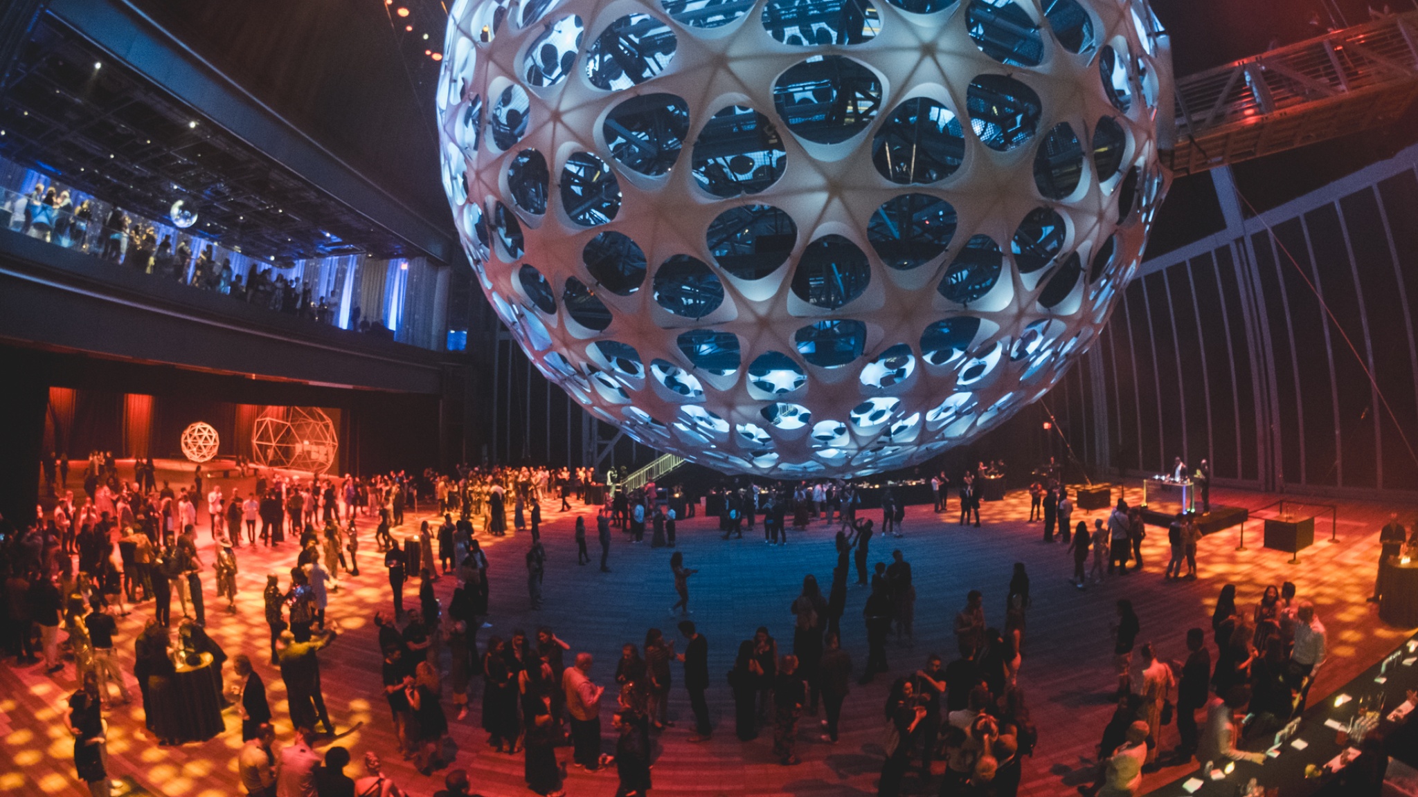 A crowd gathers beneath a large spherical concert hall suspended in air in a vast performance space. The sphere can be seen into through large apertures that make it resemble a wiffle ball. It is lit in pale blue light, and golden light illuminates the ground around its edges. 