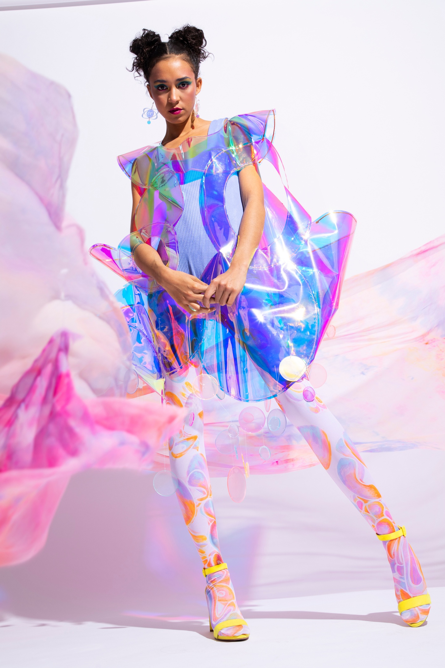 Model poses in a short, structured smock made of iridescent plastic film with rigid sleeve caps and upward-turned cuffs around the hips.