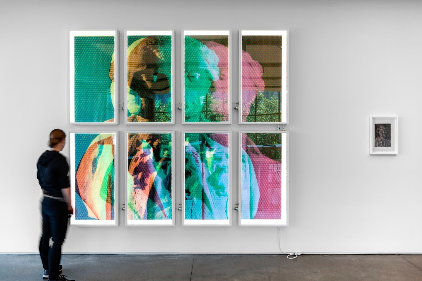 A person standing in front of an artwork made from three photographs converted into red, green, and blue monochrome tonal scales respectively, ultrachrome printed onto translucent, archival plasticized paper, cut by hand into 15mm-wide strips and triaxial woven together by hand. Backlit with eight custom-built 4000K dimmable fixtures.