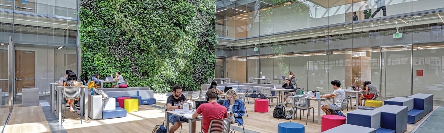 students sitting at tables inside Kuehner Court with all glass windows and a large green wall of live plants