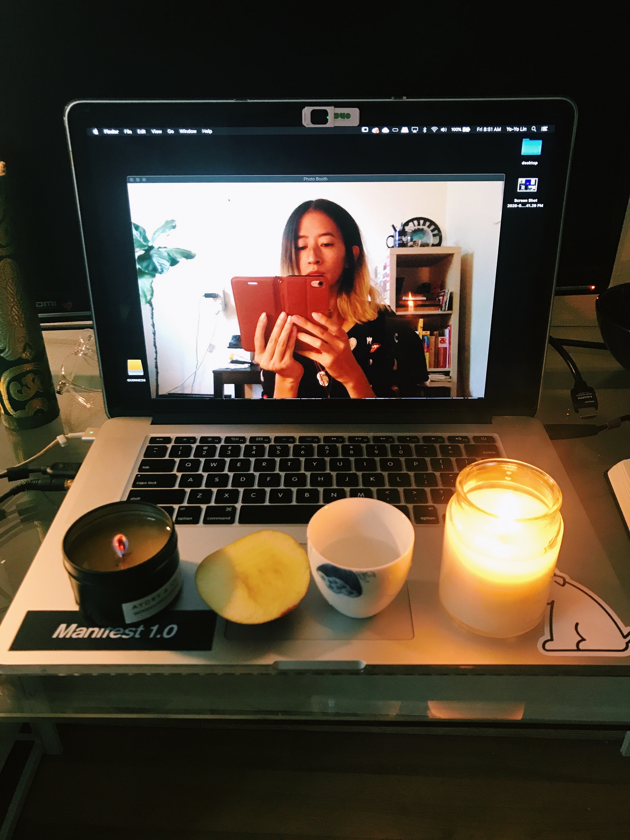 An open laptop with a webcam feed of Yo-Yo Lin in her bedroom holding her phone up to her face, obscuring her mouth. Yo-Yo is East Asian and has dark straight hair with blonde tips. On her laptop is a small altar consisting of two candles, a piece of apple and a white Chinese tea cup.