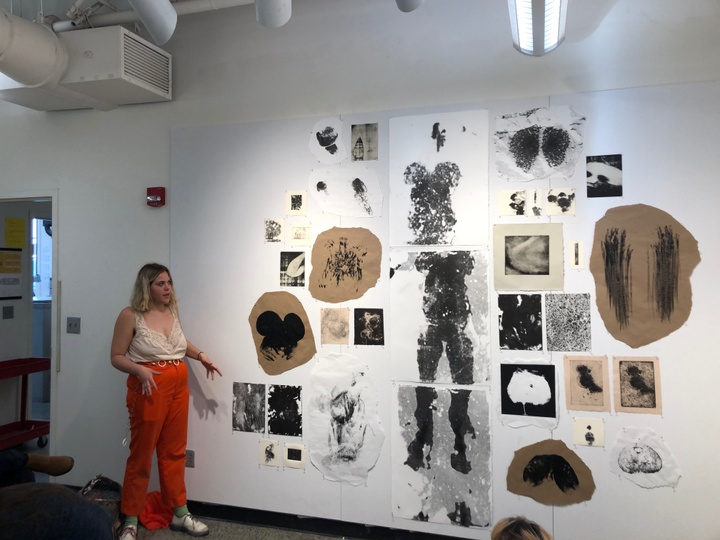 A student presents her work in class. On the wall there are black prints on various paper sizes and materials.