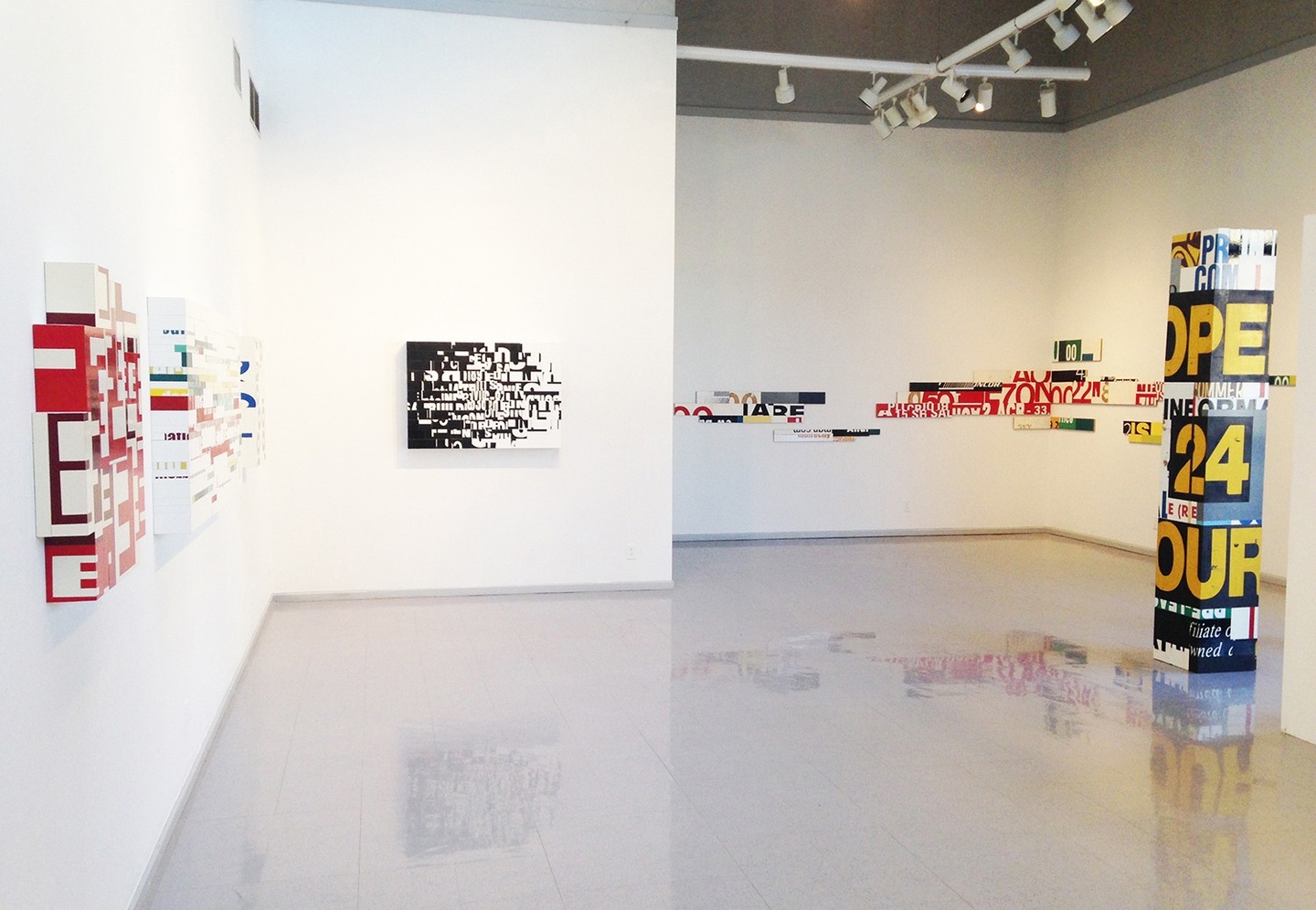 Installation view of a gallery space: on white walls several works are mounted; these art pieces are geometric sculptures, the surfaces of which are adorned with collaged/manipulated typography. In the center of this space to the right is a column/sculpture, with bold stenciled lettering.