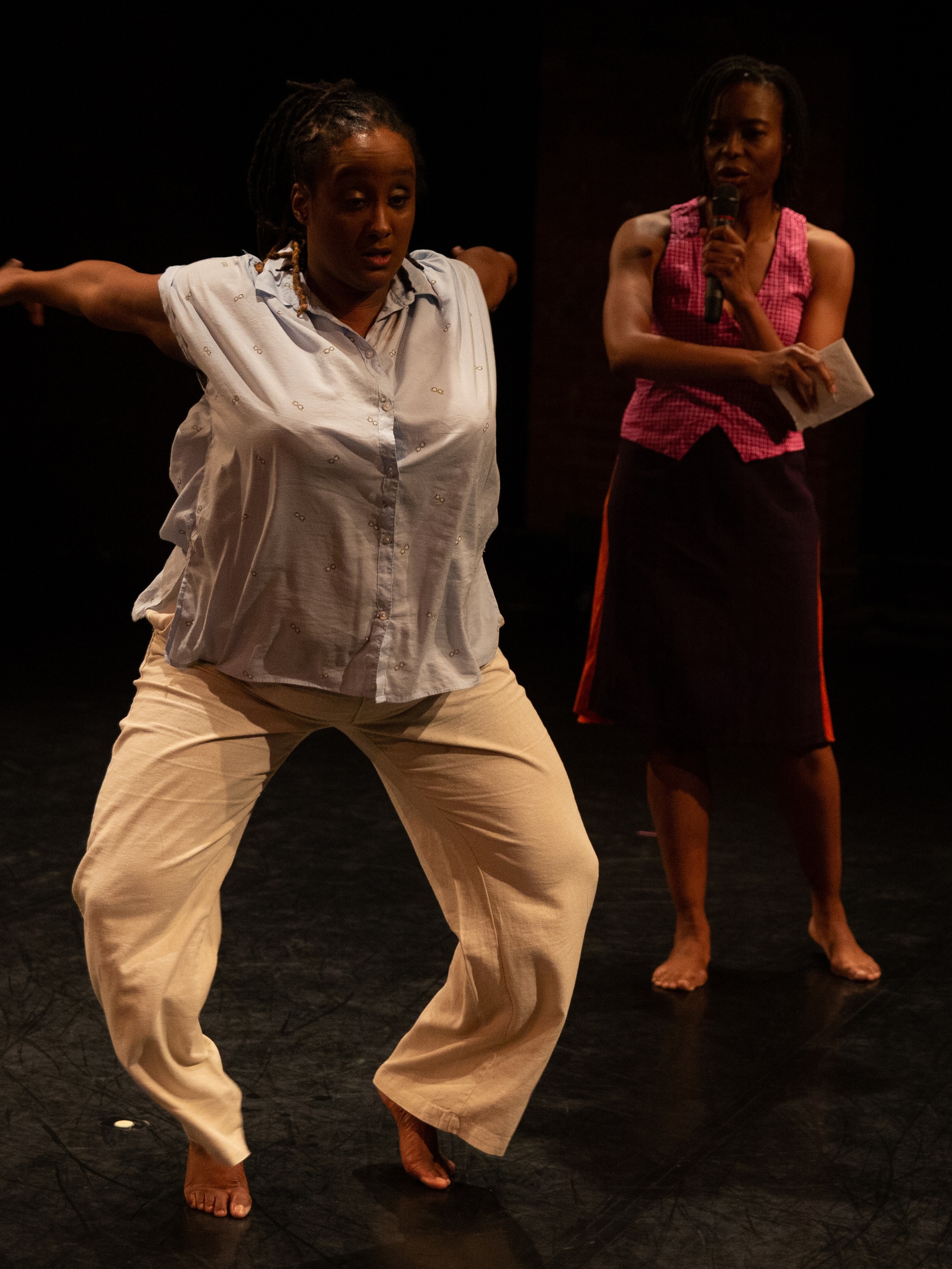 A photo of Kayla Hamilton and Nicole McClam, two dark-skinned Black women. They are both on a stage. Nicole is diagonally behind Kayla. She is wearing dark red mid-length shorts & a pink tank top. She is observing Kayla dancing. Kayla is facing front and has her knees bent as her arms are energetically swung behind her. Kayla is wearing beige pants with a white sleeveless blouse.