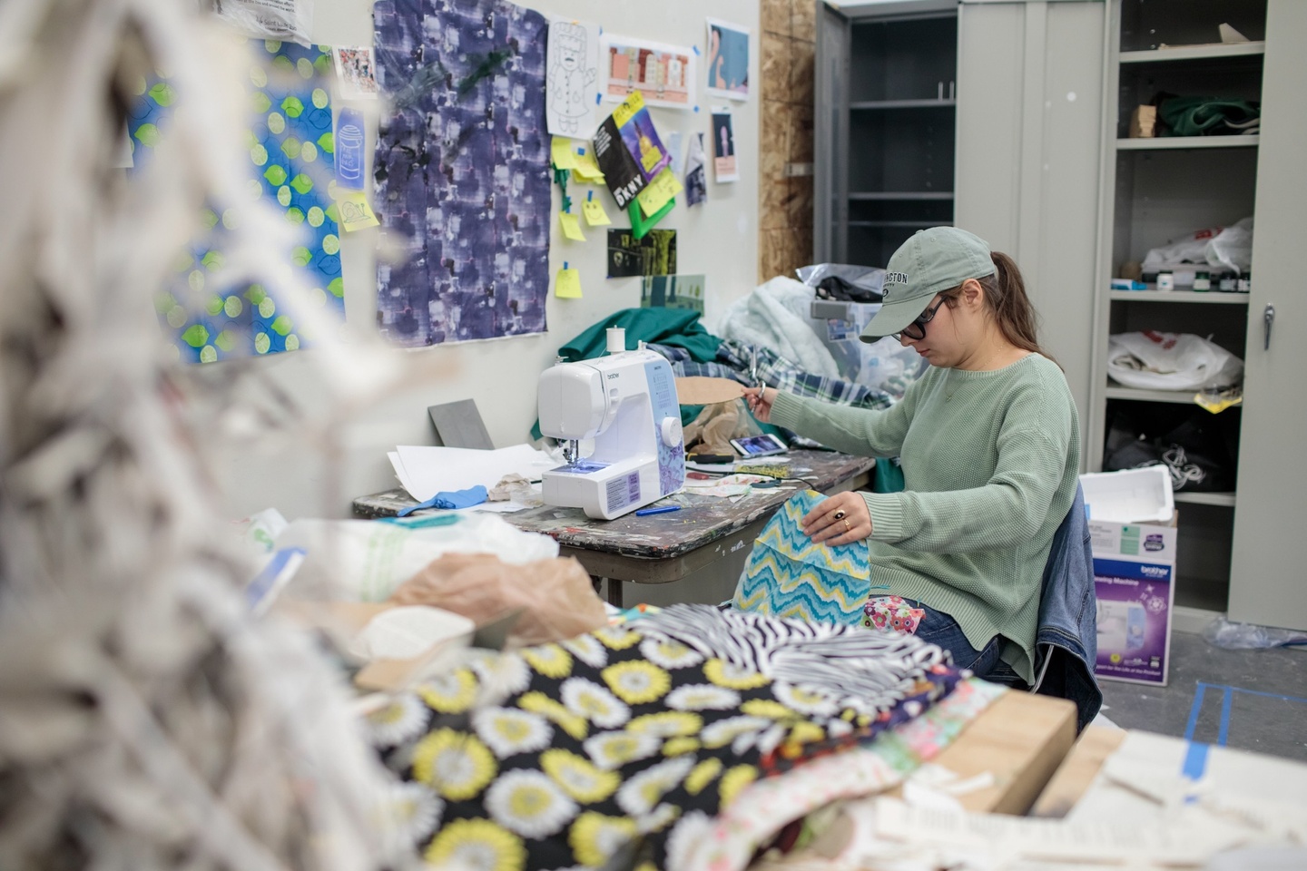 A woman sits at a sewing machine in the fashion studio. There are piles of fabric surrounding her.