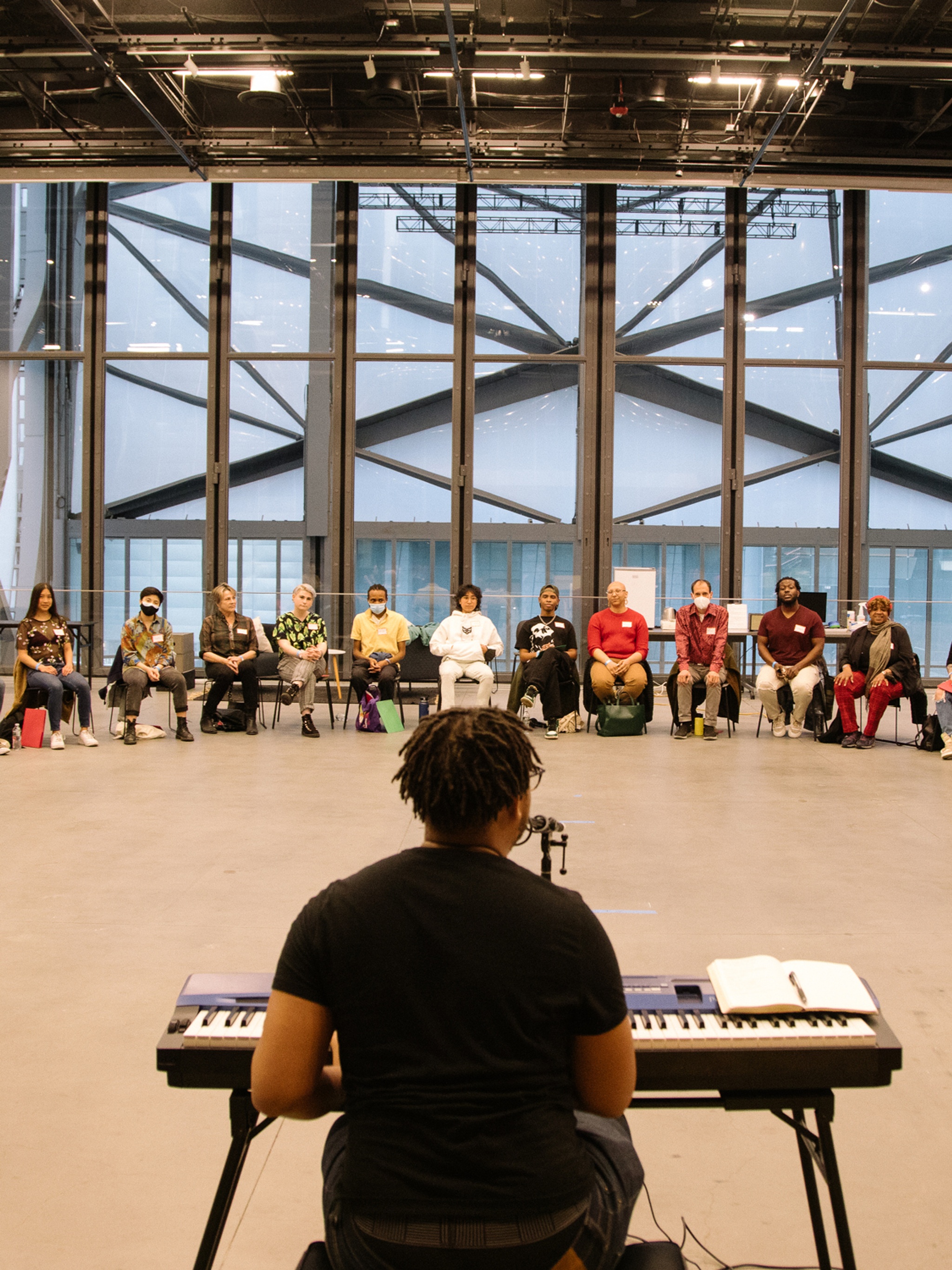 The artist Troy Anthony sits at a keyboard with his back to the camera facing a semicircle of people of various ages and races seated across a wide performance space with high ceilings at The Shed