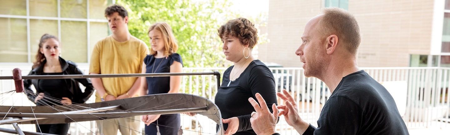 A male professor talks with his hands while teaching a group of students who are standing and looking at a sculpture