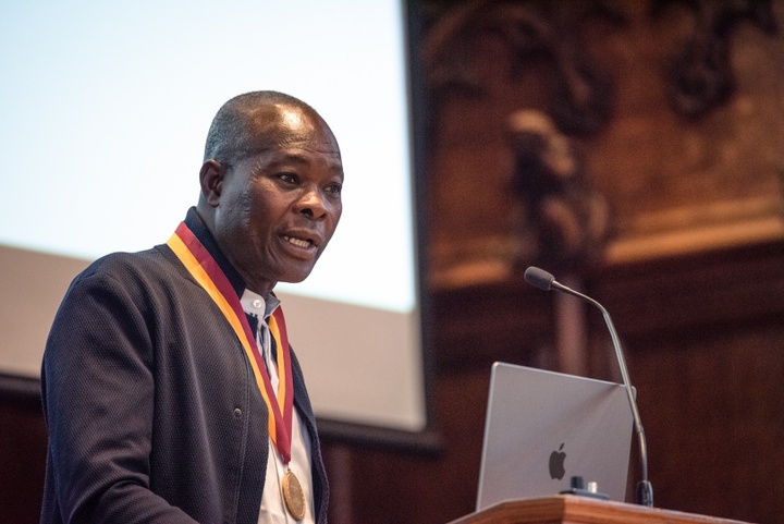 Francis Kéré, wearing a medal on a red and yellow ribbon, speaks at a podium in front of a projector screen in Graham Chapel.
