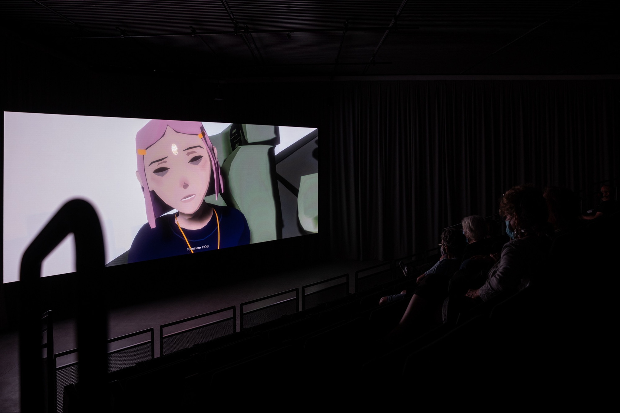 A movie screen seen over the edge of a bank of cinema seats. On the screen is a close up of an anime character's face. She has pink hair.
