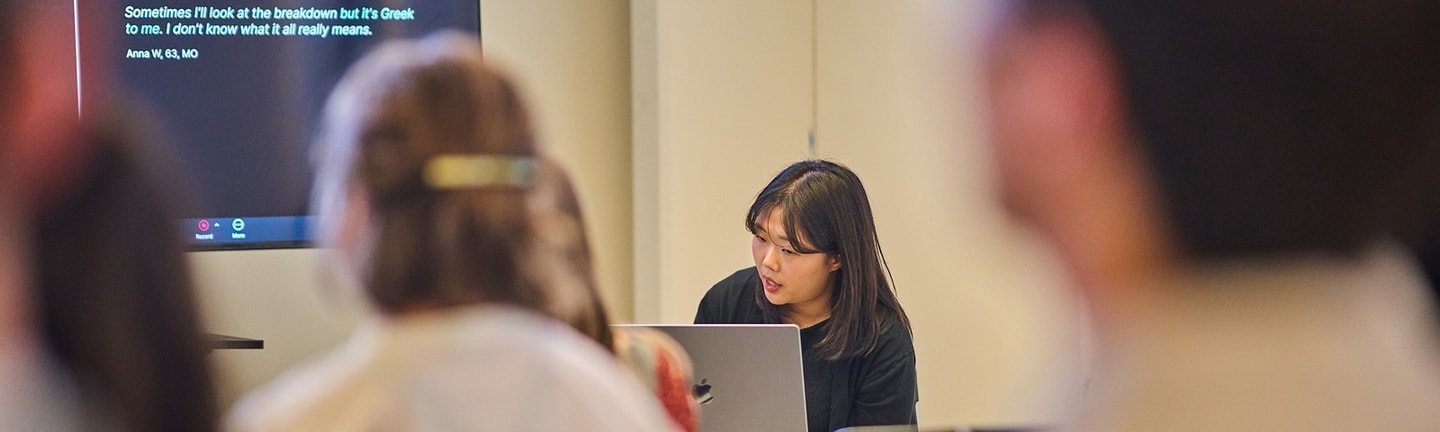 A student sits at a computer while presenting her project to a group of people seated.